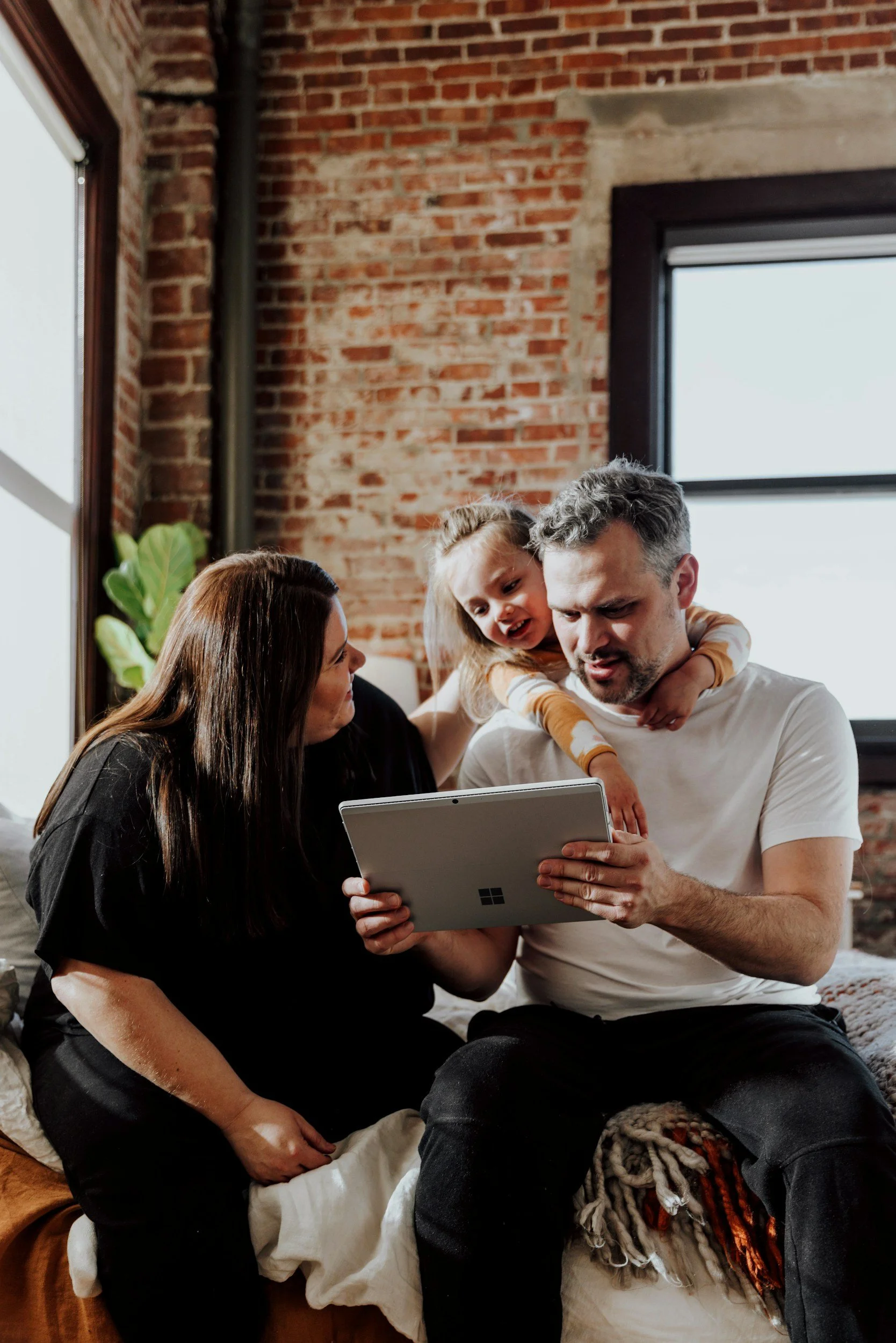 Family of three gathered around a tablet inside a room with exposed brick wall, large window, and a plant in the background.