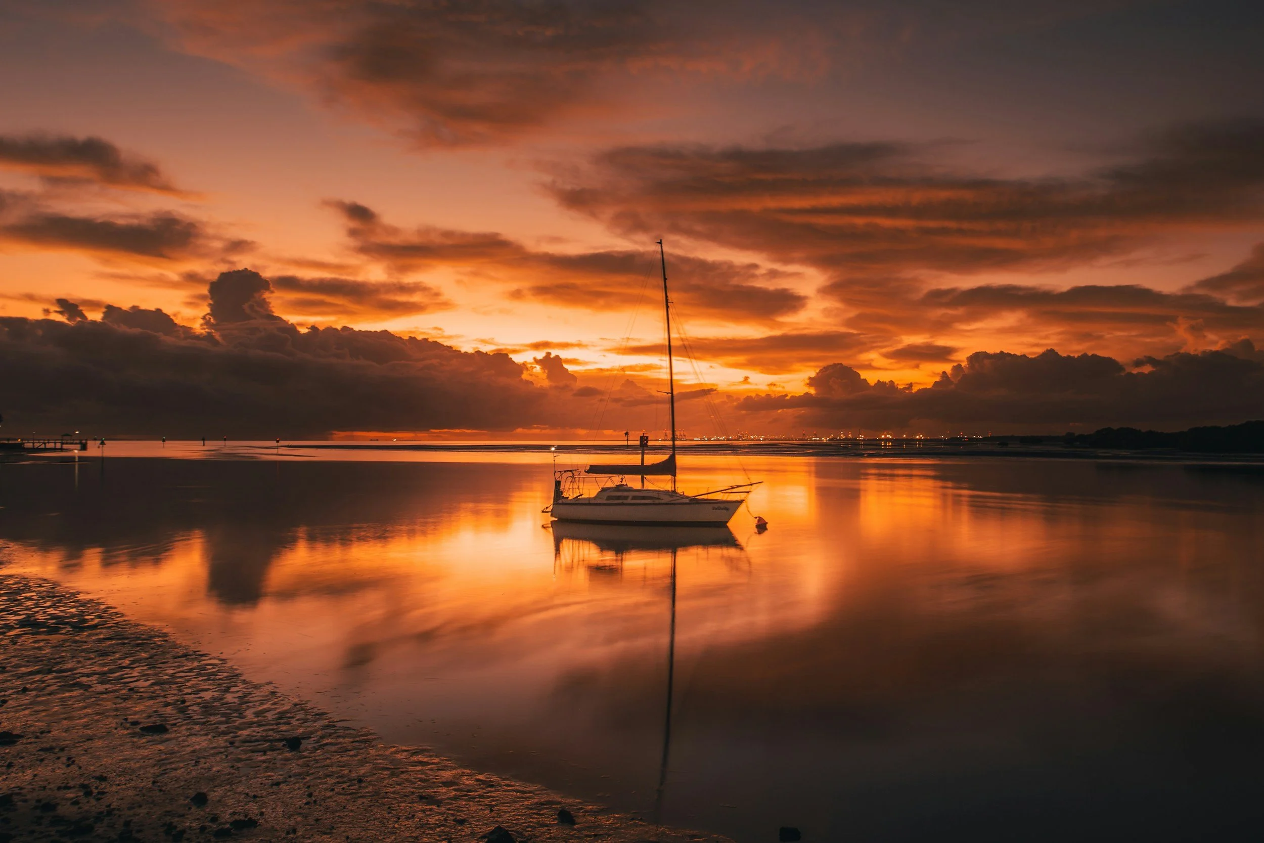 A sailboat floating on calm water during a vibrant orange and purple sunset with clouds in the sky.