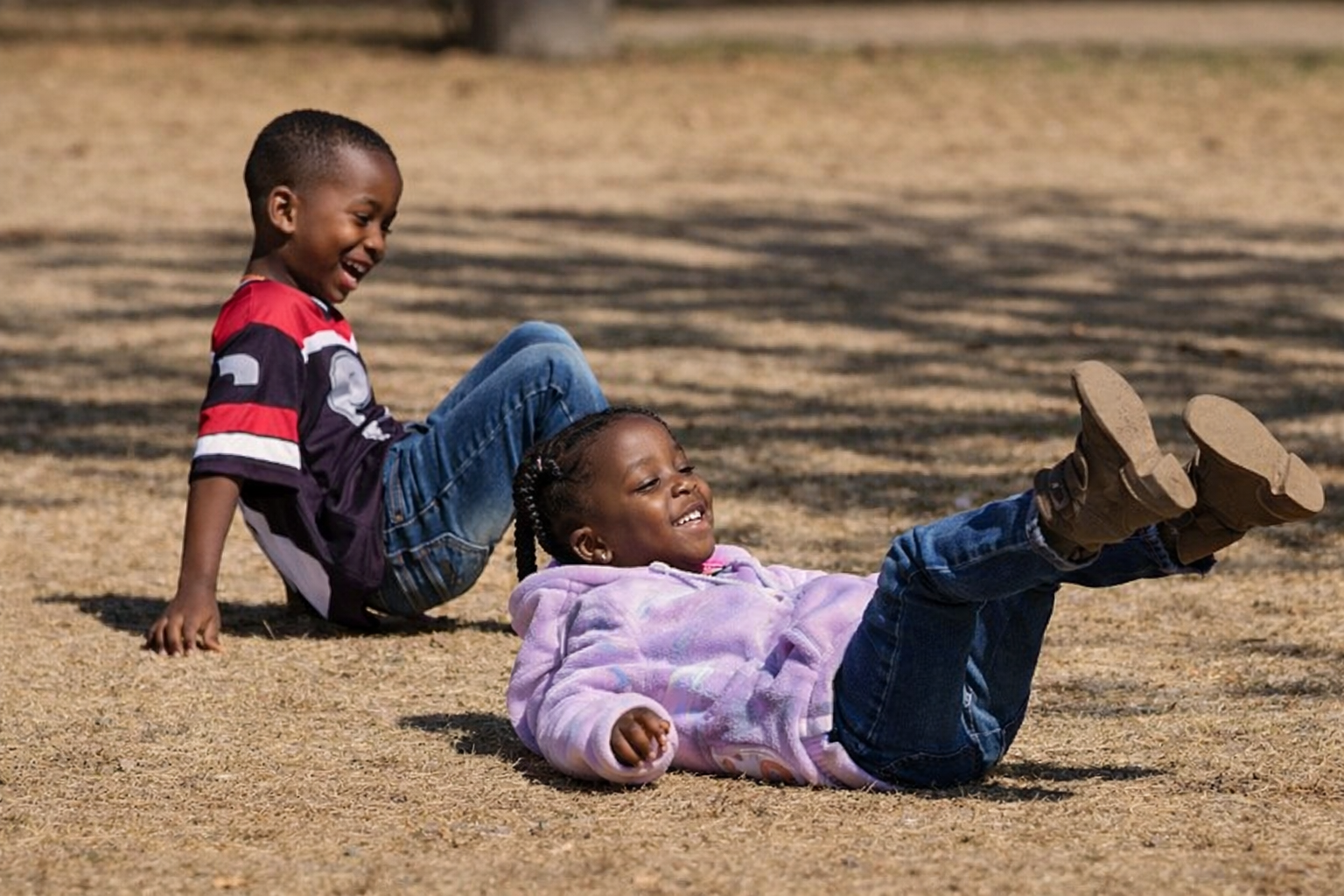 Two children, a boy and a girl, playing on the grass outdoors, smiling and laughing, with the boy sitting behind the girl who is lying on her back.