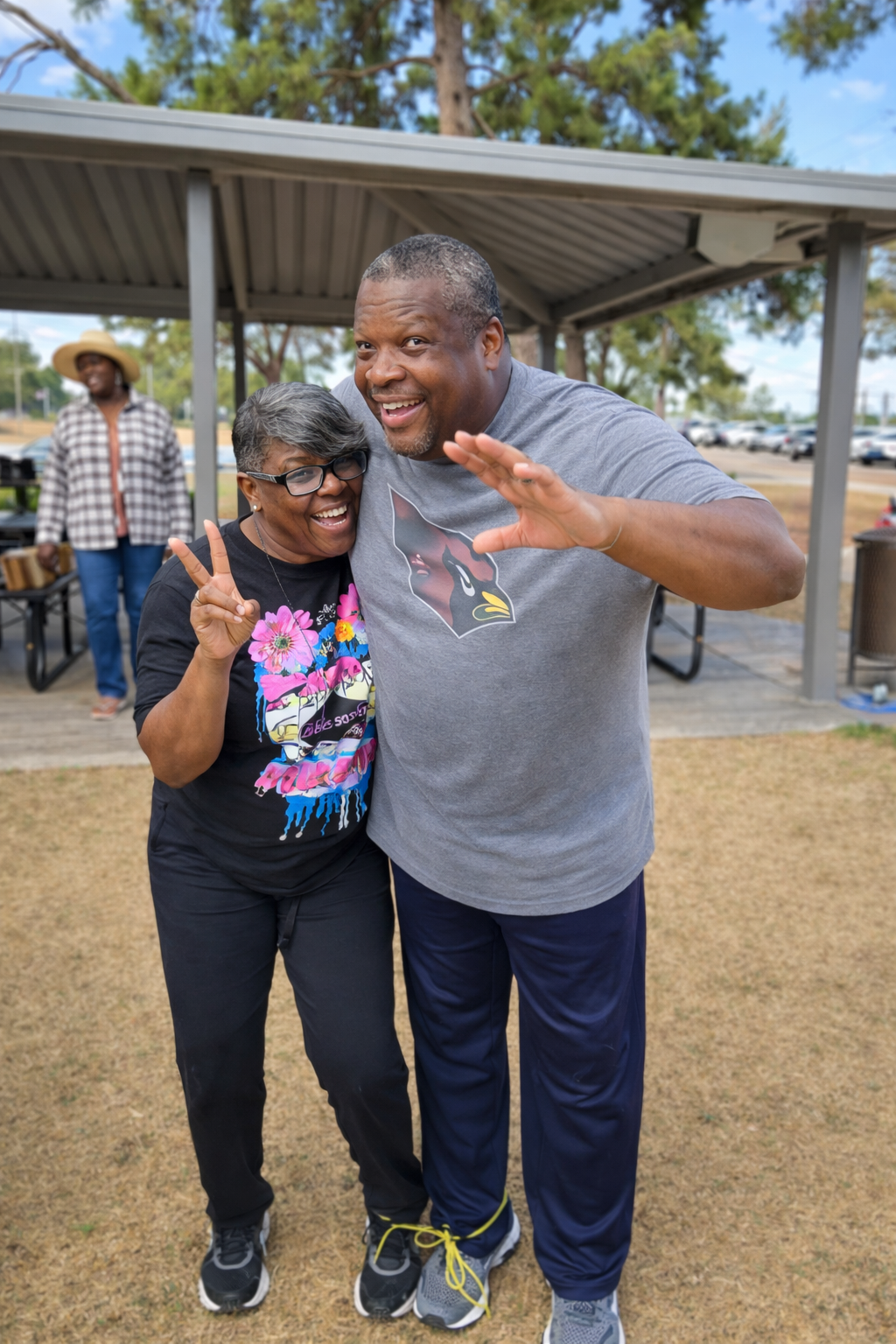 Two people, an older woman with glasses and a man, smiling and posing outdoors, making peace signs and waving, with a woman in a straw hat standing in the background near a pavilion.