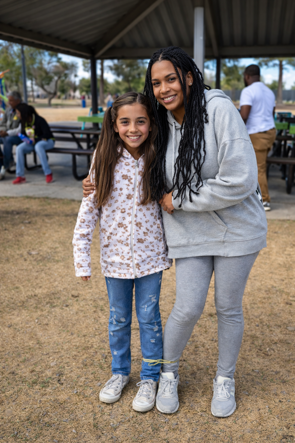 A woman and a young girl smiling and standing close together outdoors under a pavilion. The woman wearing a gray hoodie and sweatpants, the girl in a animal print hoodie and distressed jeans. Other people are in the background sitting on benches.