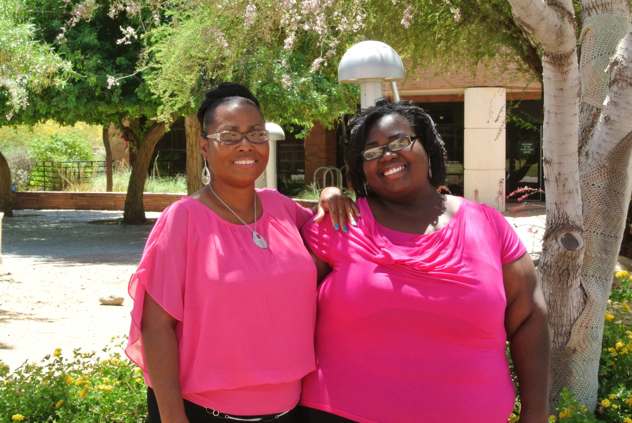 Two women wearing pink tops standing outdoors under a tree, smiling at the camera.