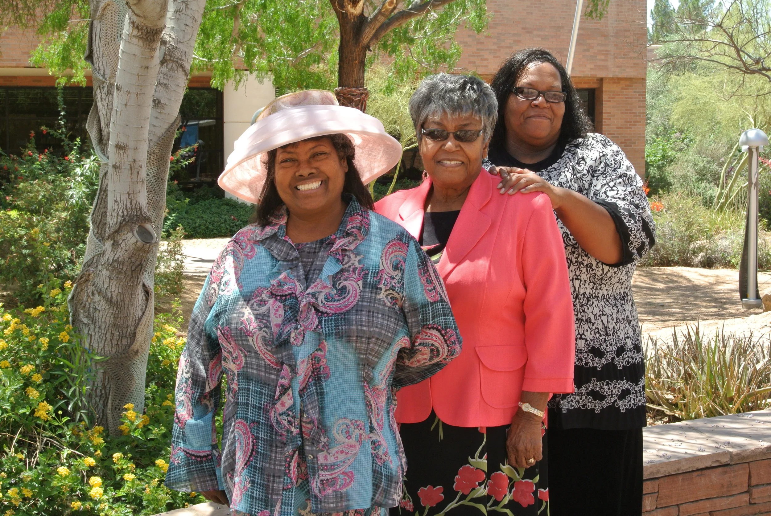 Three women smiling outdoors in a garden, with trees and a brick building in the background. The woman in front is wearing a colorful patterned blouse and a pink hat. The woman in the middle is dressed in a pink blazer and a black dress with floral p