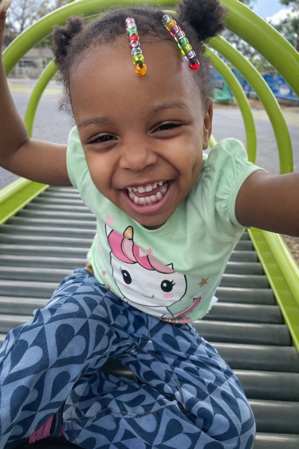 A smiling young girl with beaded hair, wearing a pastel green unicorn t-shirt and blue patterned pants, is sitting on a playground slide and leaning towards the camera, enjoying her time outside.