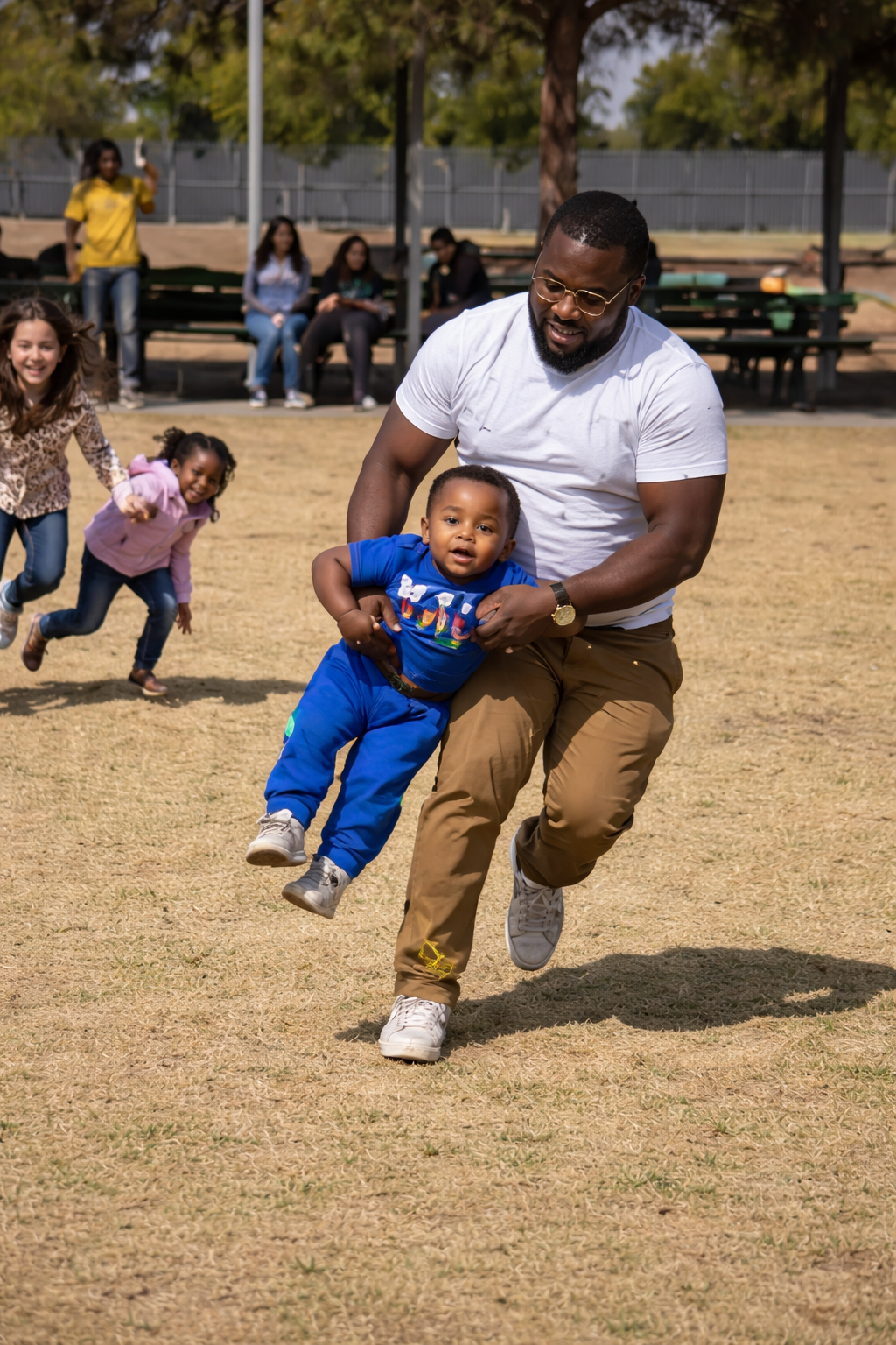 A man in a white t-shirt and brown pants is playfully lifting a young boy in a blue shirt and pants at a park, with children and adults in the background.