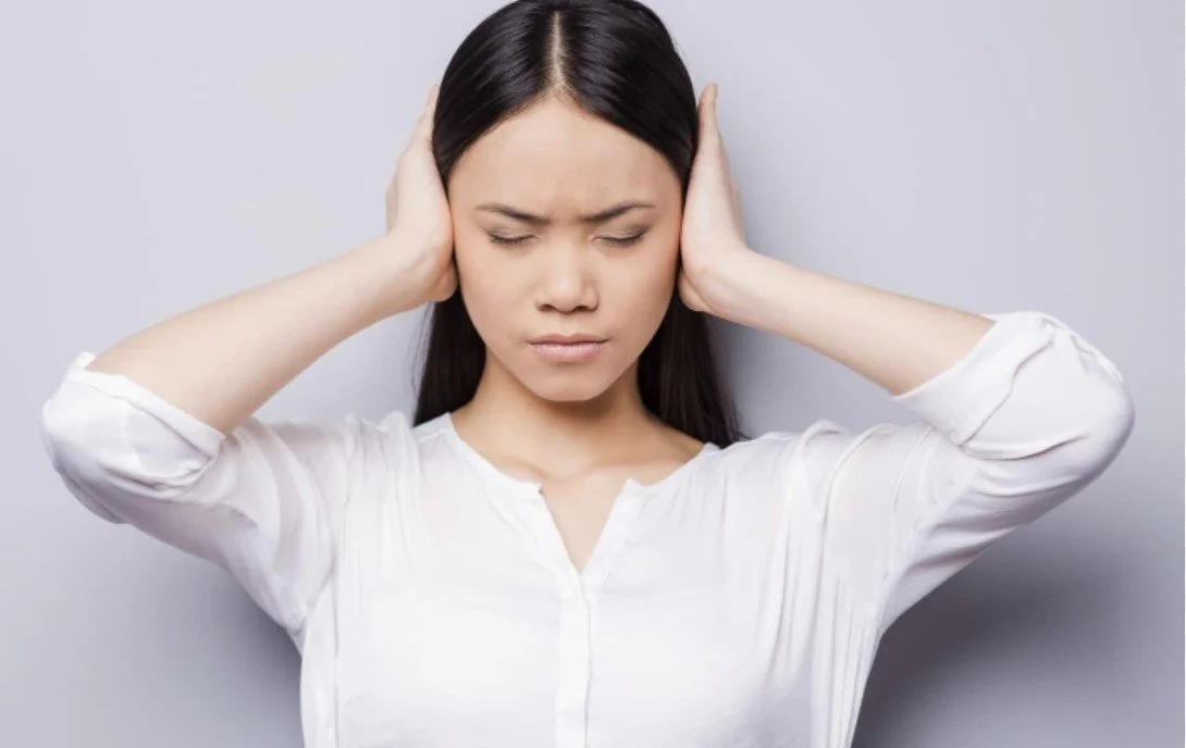 A woman with dark hair wearing a white shirt, holding her head with both hands and appears to be experiencing a headache or stress.
