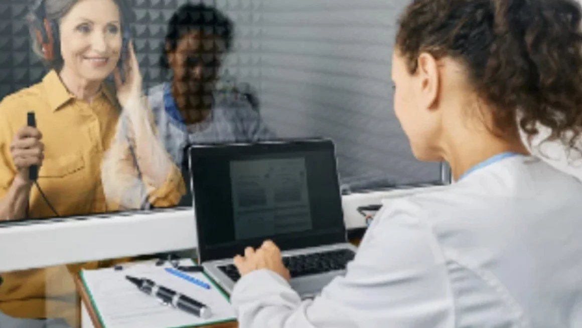 A woman in a yellow shirt wearing headphones holds a microphone inside soundproof glass, with a woman and a man in the background, and another woman with curly hair working on a laptop at a desk.
