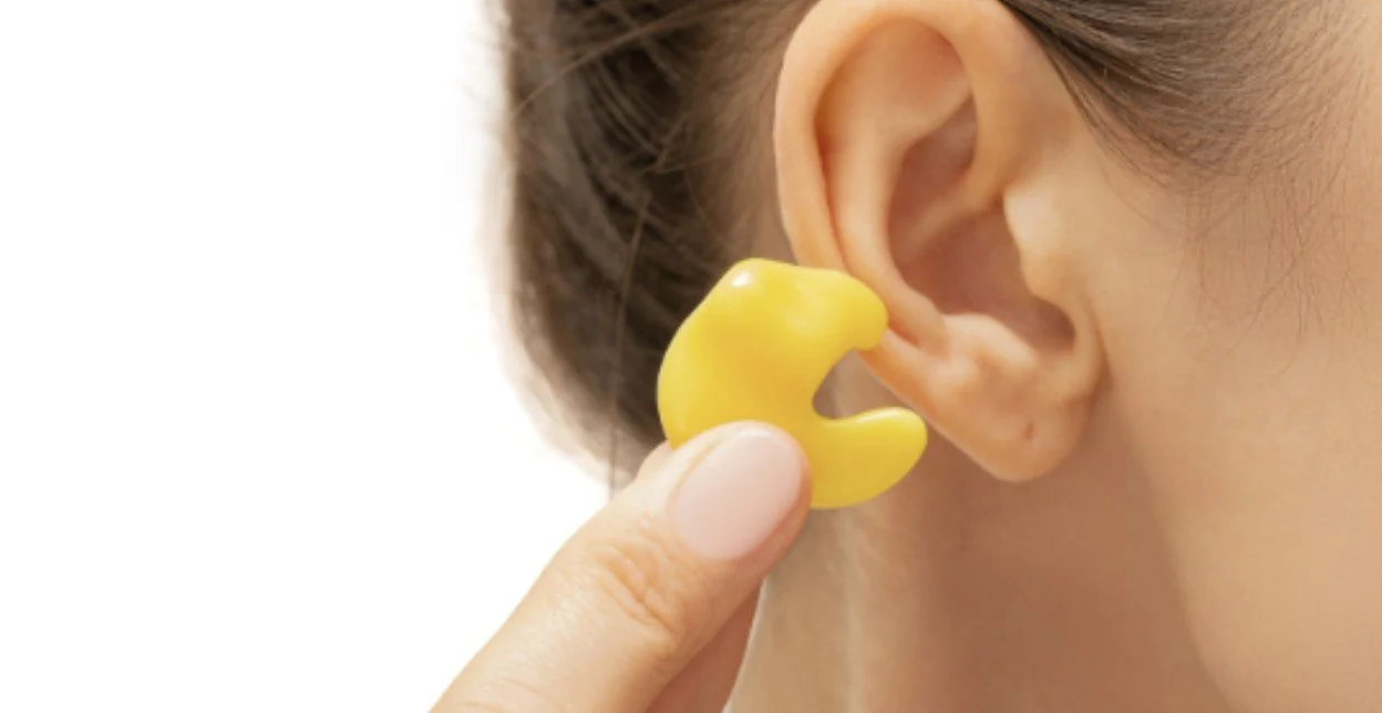 A woman inserting a yellow earplug into her ear for hearing protection.
