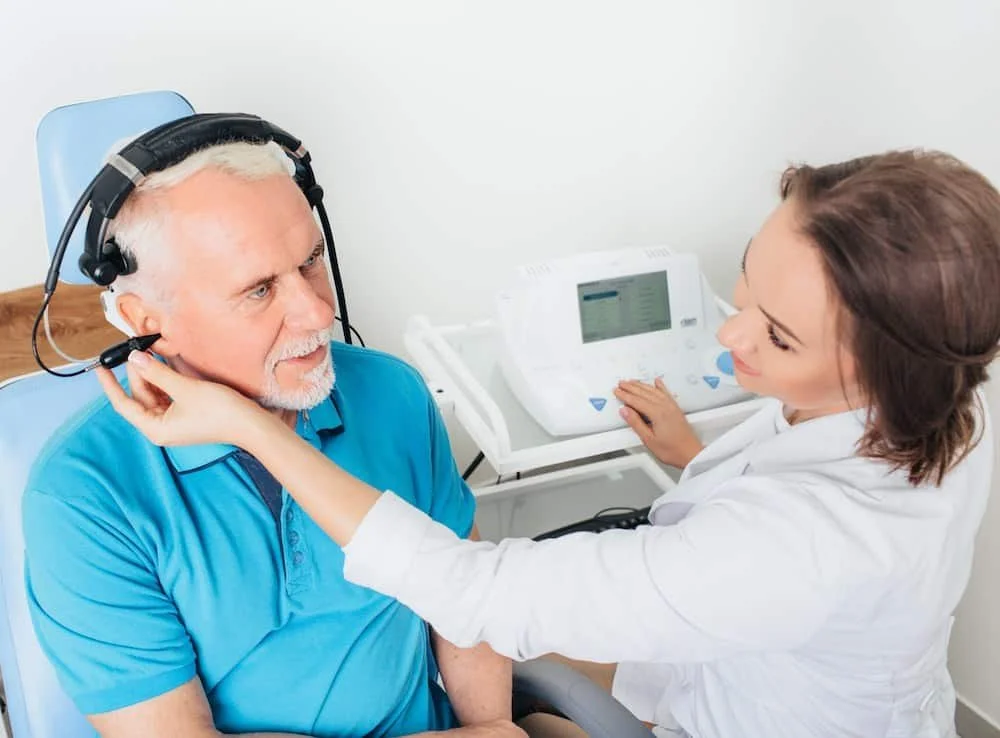 Doctor placing a hearing aid on an older man who is wearing a headset, in a medical examination room.