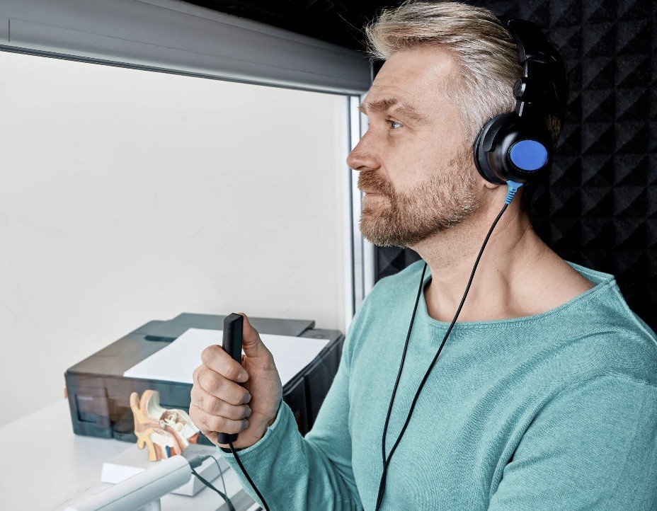 A man with gray hair, beard, and blue headphones sitting in a soundproof booth, holding a microphone in his right hand, with a model of a human ear on his desk.