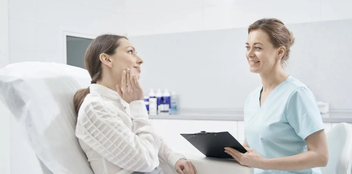 A woman in a hospital bed talking to a nurse or doctor who holds a clipboard in a clinical setting.