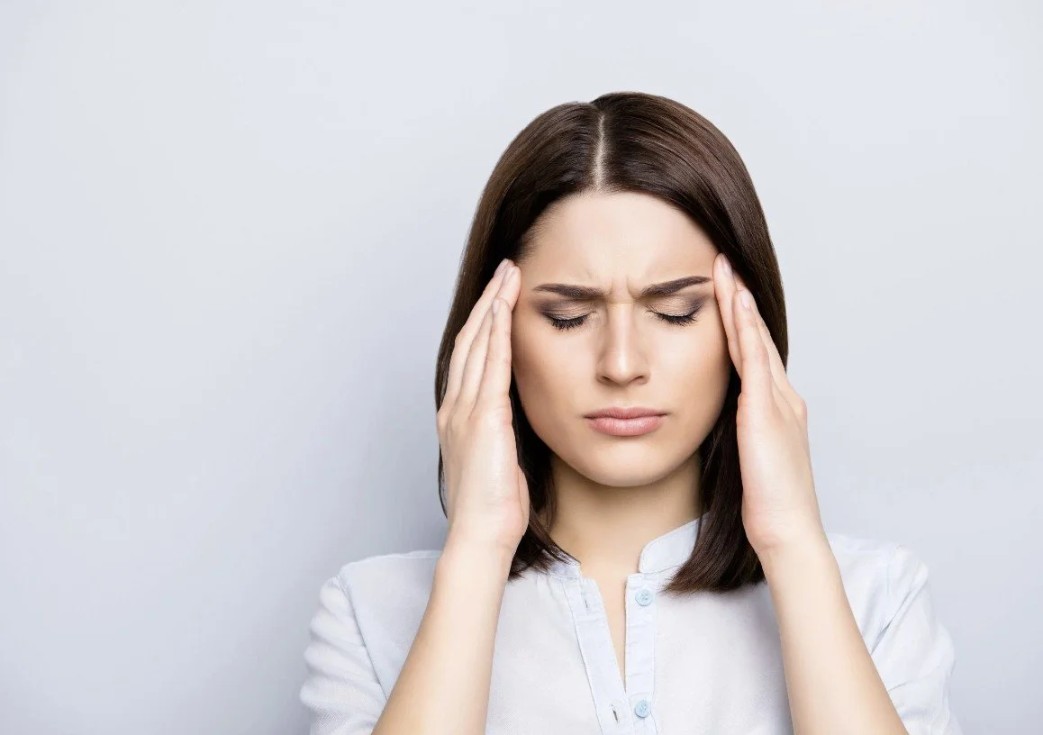 A woman with brown hair holding her head with her eyes closed, appearing to have a headache or feeling stressed.