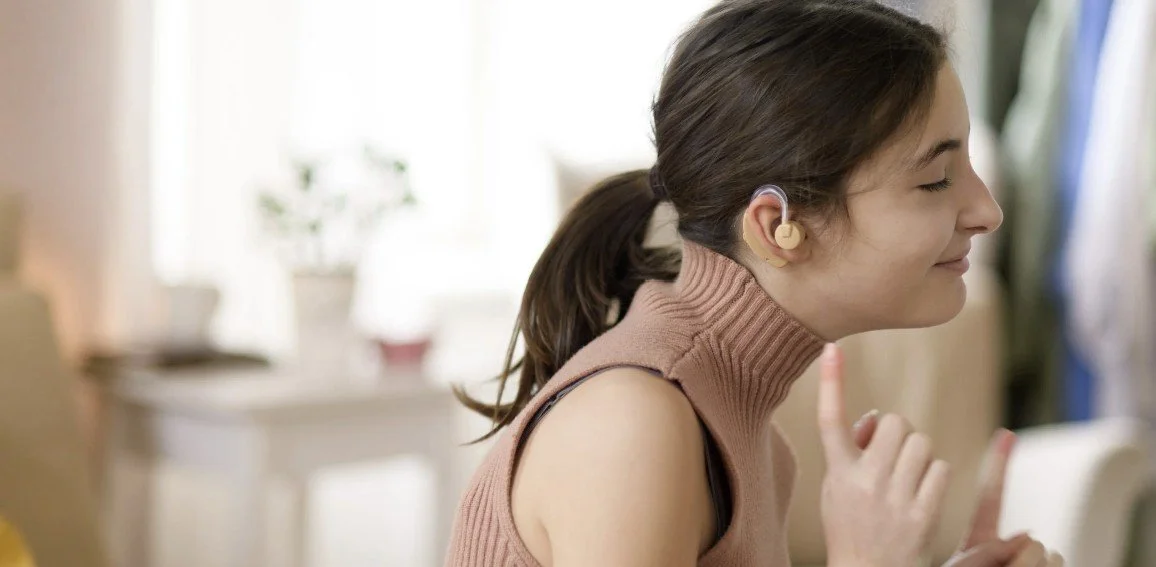 A woman with brown hair in a ponytail wearing a sleeveless turtleneck sweater, smiling with eyes closed, listening to music with wireless earbuds in a bright room.