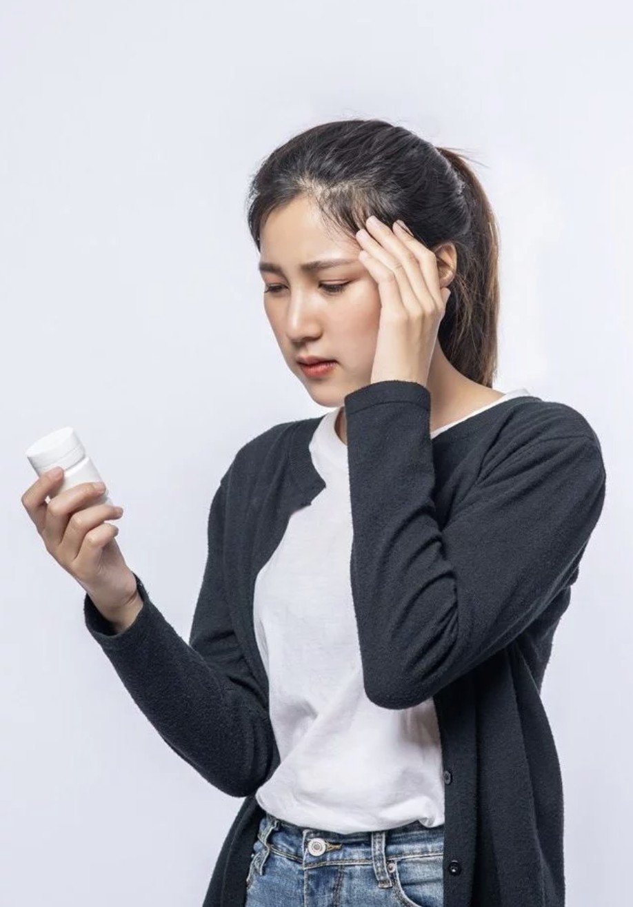 A young woman looks concerned as she holds a bottle of pills in one hand and touches her forehead with her other hand.