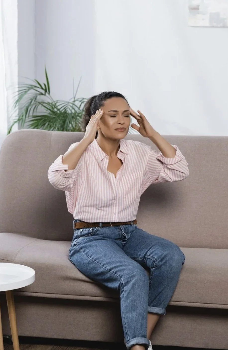 A woman sitting on a beige couch, holding her head with a pained expression, experiencing a headache or stress, with a green plant and white wall in the background.