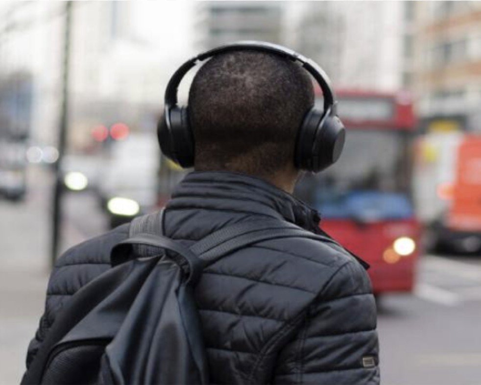 Person with short hair wearing large headphones and a black puffer jacket, carrying a backpack, standing on a city street near a red bus.