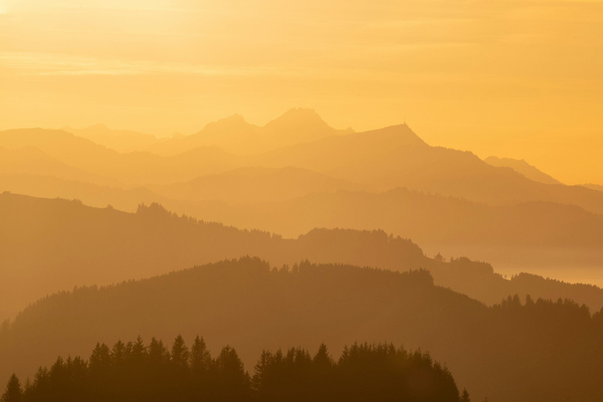 Paysage de montagnes avec plusieurs couches de collines couvertes d'arbres, sous un ciel orange au lever ou coucher du soleil.