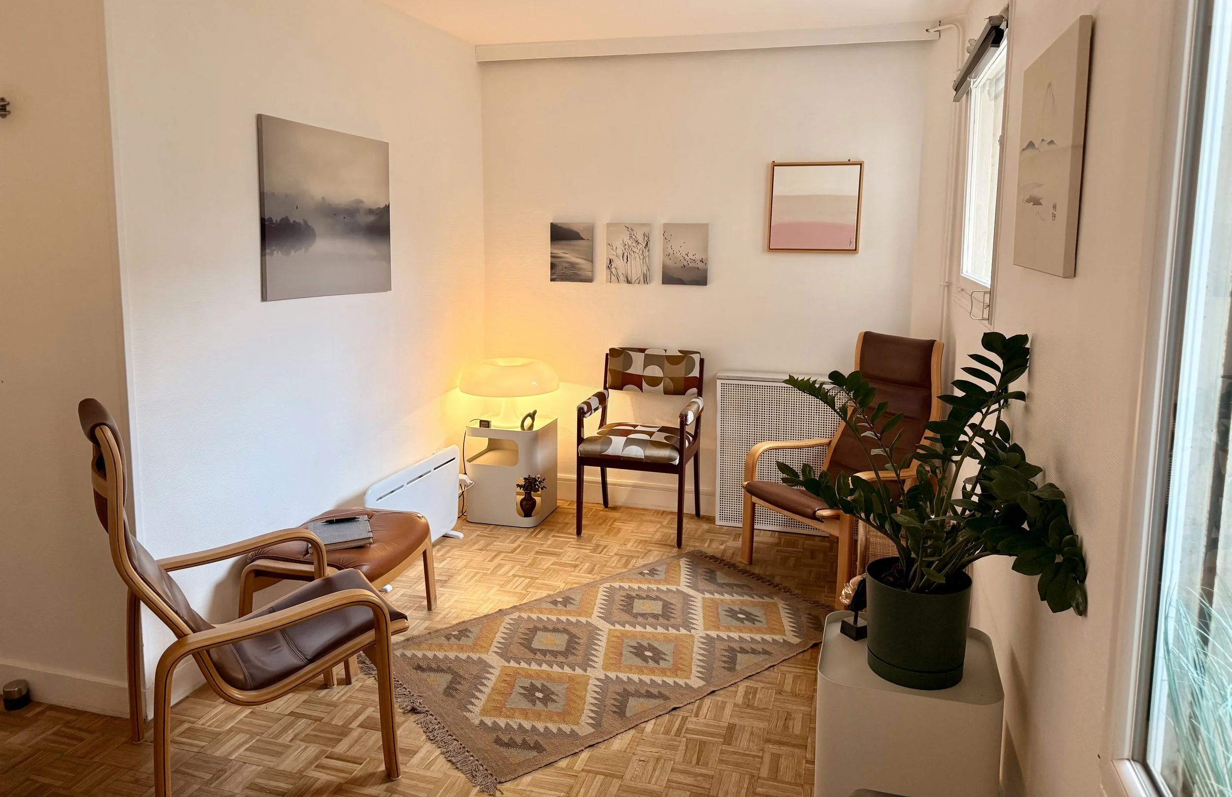 Psychanalyste Bordeaux Salon cosy avec fauteuils en bois, tapis géométrique, plantes, lampadaire, tableaux abstraits, fenêtre laissant entrer la lumière naturelle.