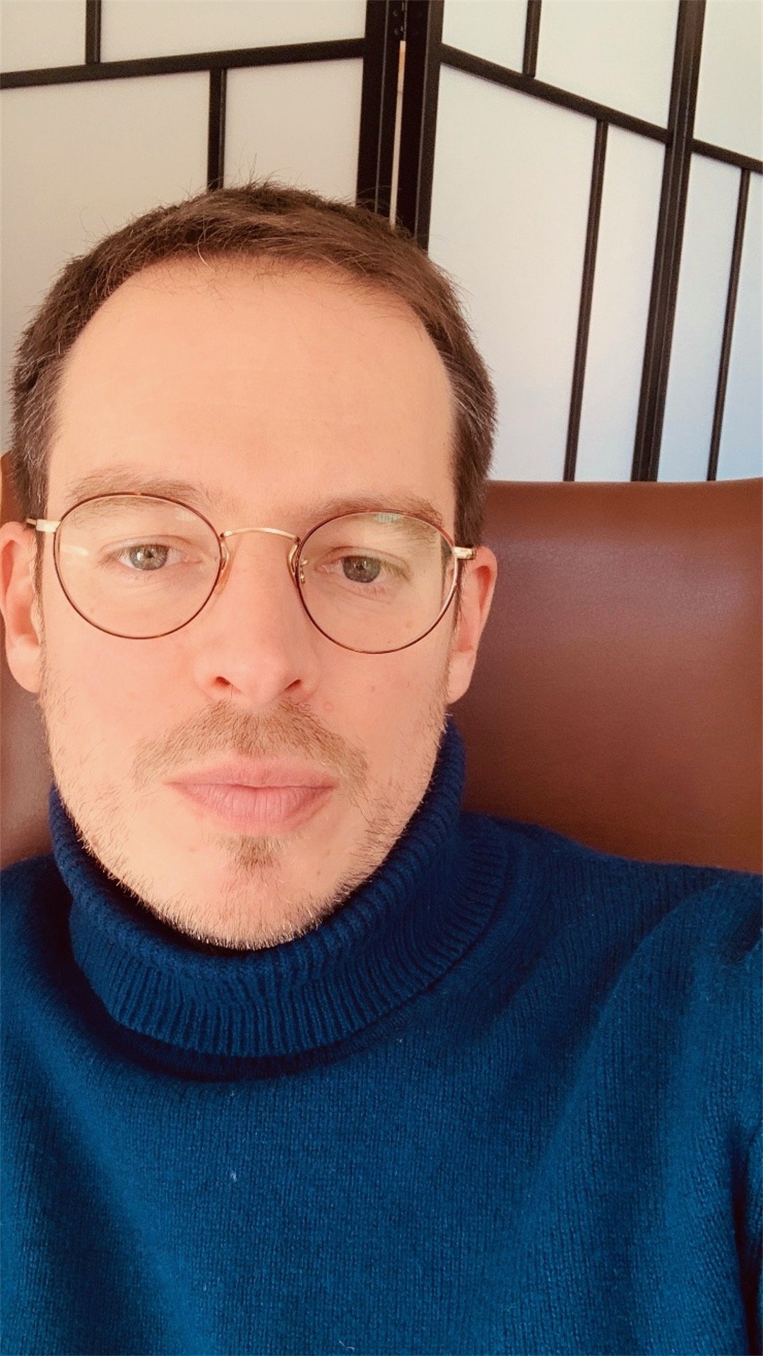 Psychanalyste un homme portant des lunettes rondes, un pull bleu, assis dans un lieu avec un décor en papier japonais