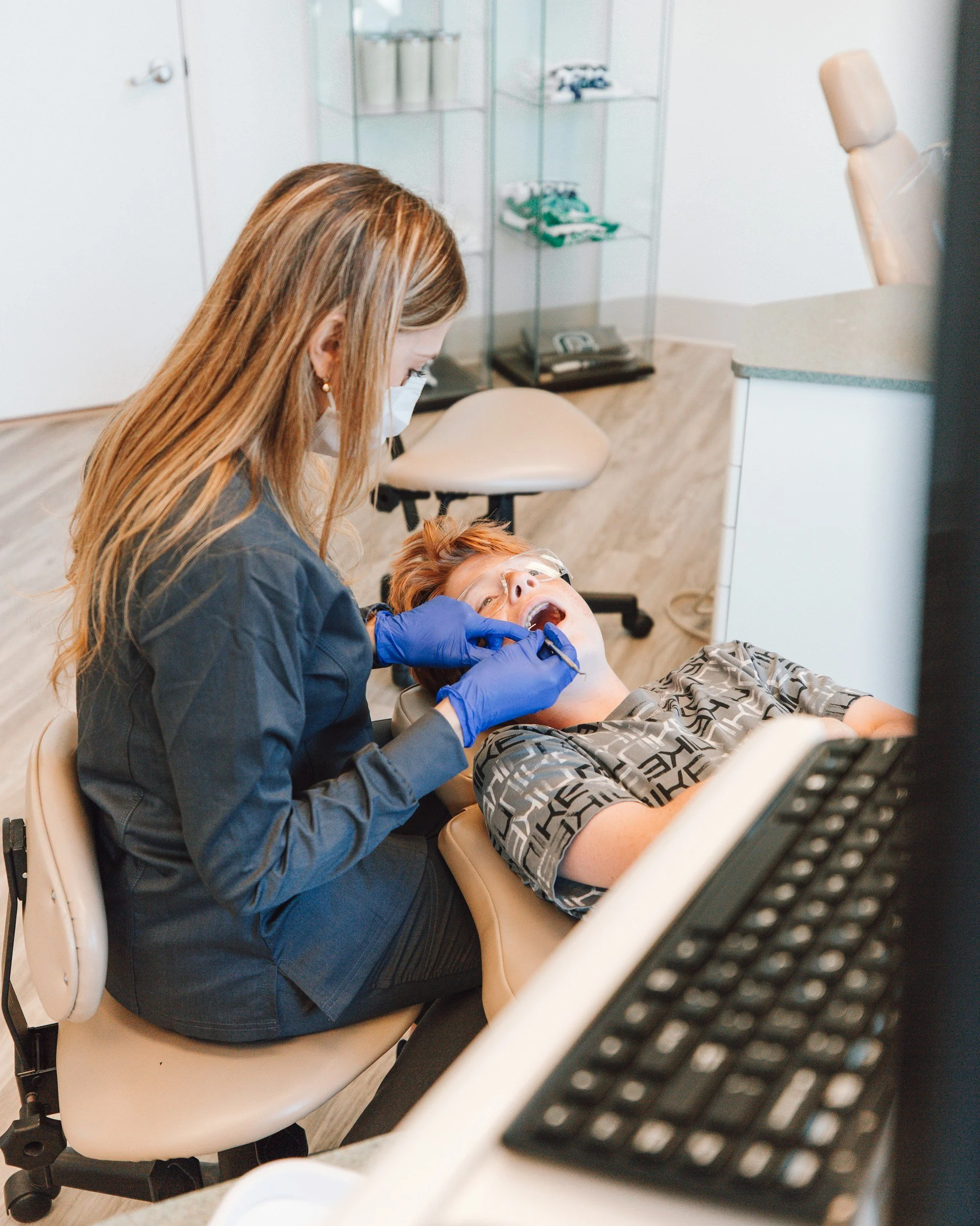 A woman with long hair and a face mask helps a young patient with orange hair and glasses who is lying on a dental chair, with her mouth open, receiving dental treatment in a clinic.