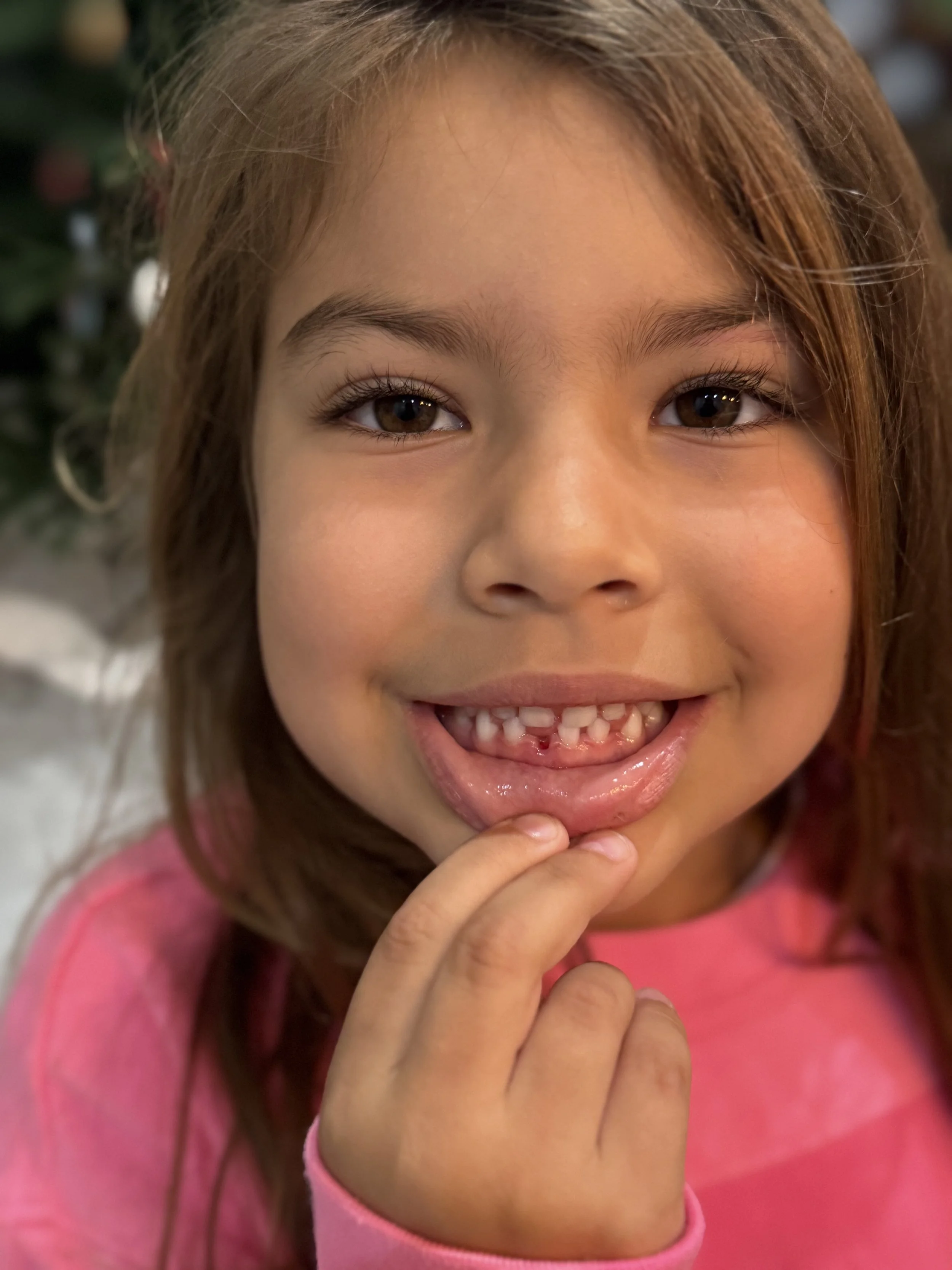 A young girl with brown hair and brown eyes smiling and touching her chin with her fingers, wearing a pink shirt.