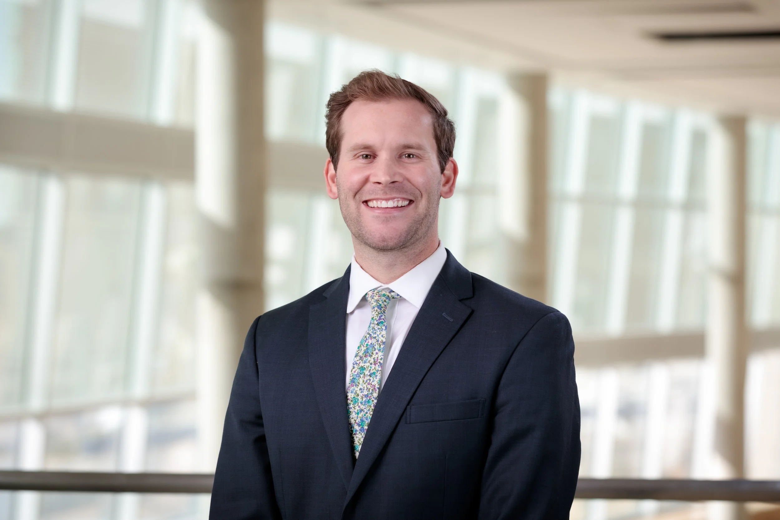 A smiling man in a dark suit with a white shirt and floral tie standing indoors with large windows and modern architecture in the background.