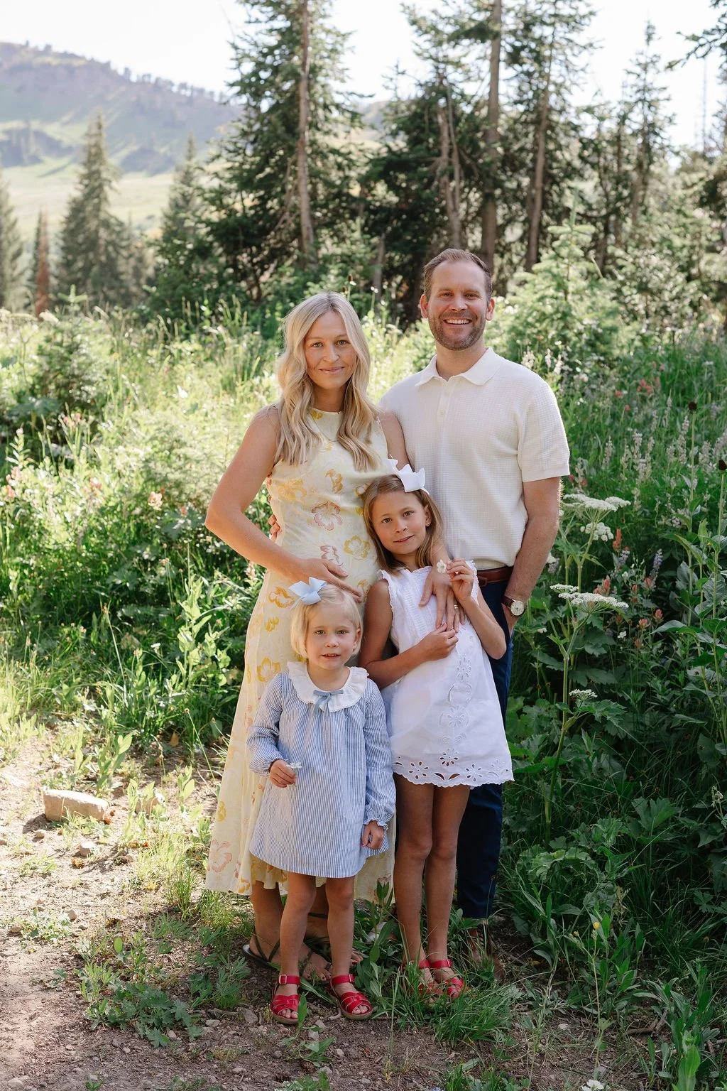 A family of five smiling outdoors in a green, forested area with mountains in the background. The mother is pregnant, wearing a yellow dress, and the father is in a white shirt. Two young girls are in front, dressed in light-colored dresses with bows in their hair.