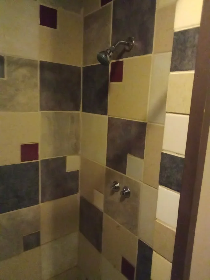 Corner of a shower with multi-colored tile walls, a shower head, and two control knobs.