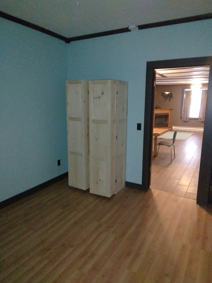 Empty room with light blue walls, hardwood floor, black trim, and a wooden closet in the corner. An adjacent room is visible through an open doorway, showing a dining area with a table, chair, and a window.