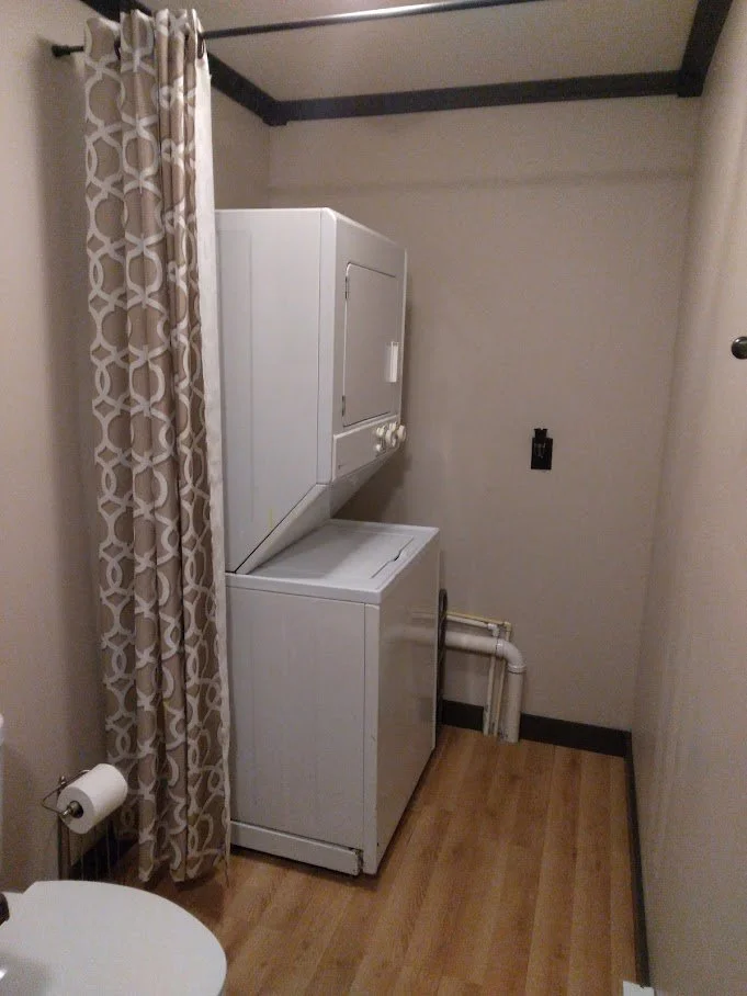 A laundry room with a stacked washer and dryer, a curtain on the left, a toilet in the corner, and exposed pipes in the back.