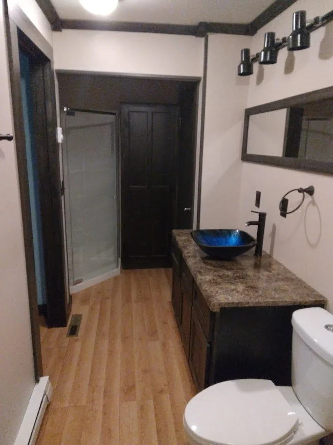 Small bathroom with hardwood floor, black door cabinets, granite countertop, black vessel sink, wall-mounted mirror, black light fixtures, toilet, and standing shower with glass door.