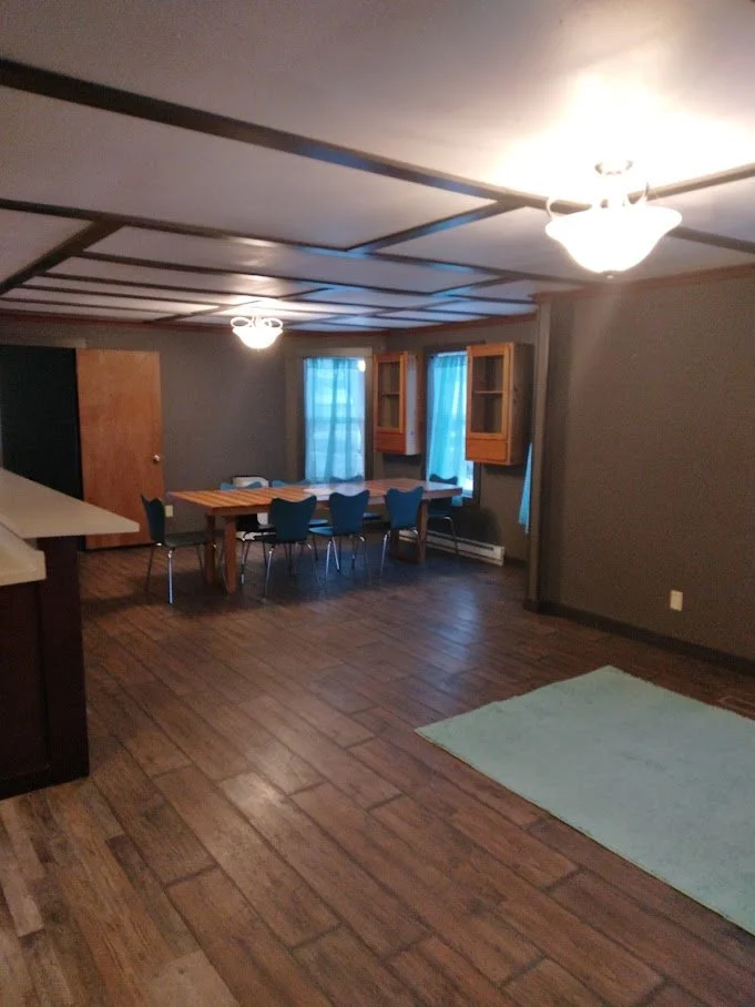 A dining area with a wooden table and six blue chairs in a room with dark gray walls, wood-look flooring, a light-colored rug, and three windows with blue curtains. The ceiling has a decorative grid pattern and a ceiling fan with a light.