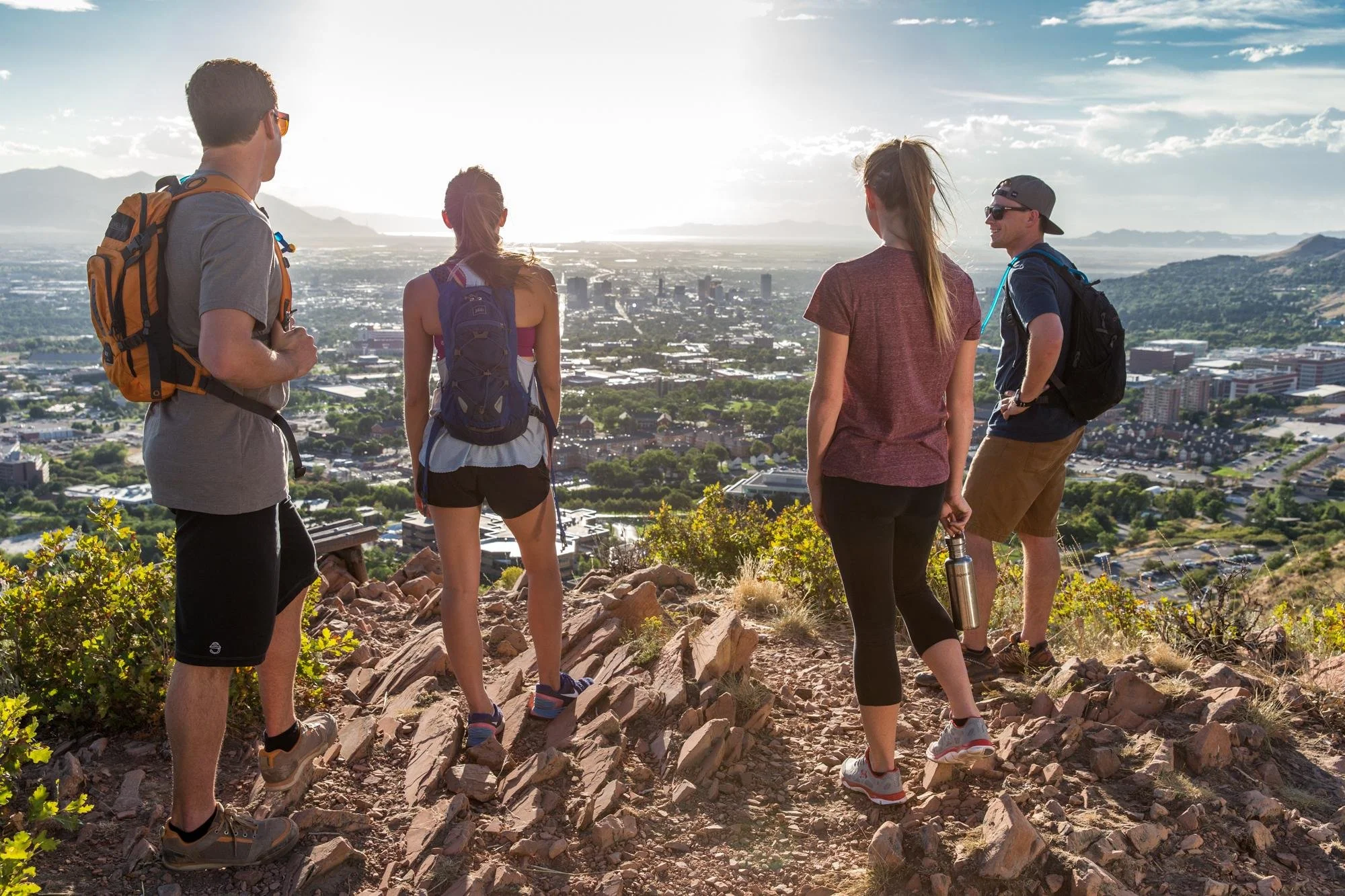 Five hikers standing on a rocky trail overlooking Salt Lake City and the surrounding mountain.