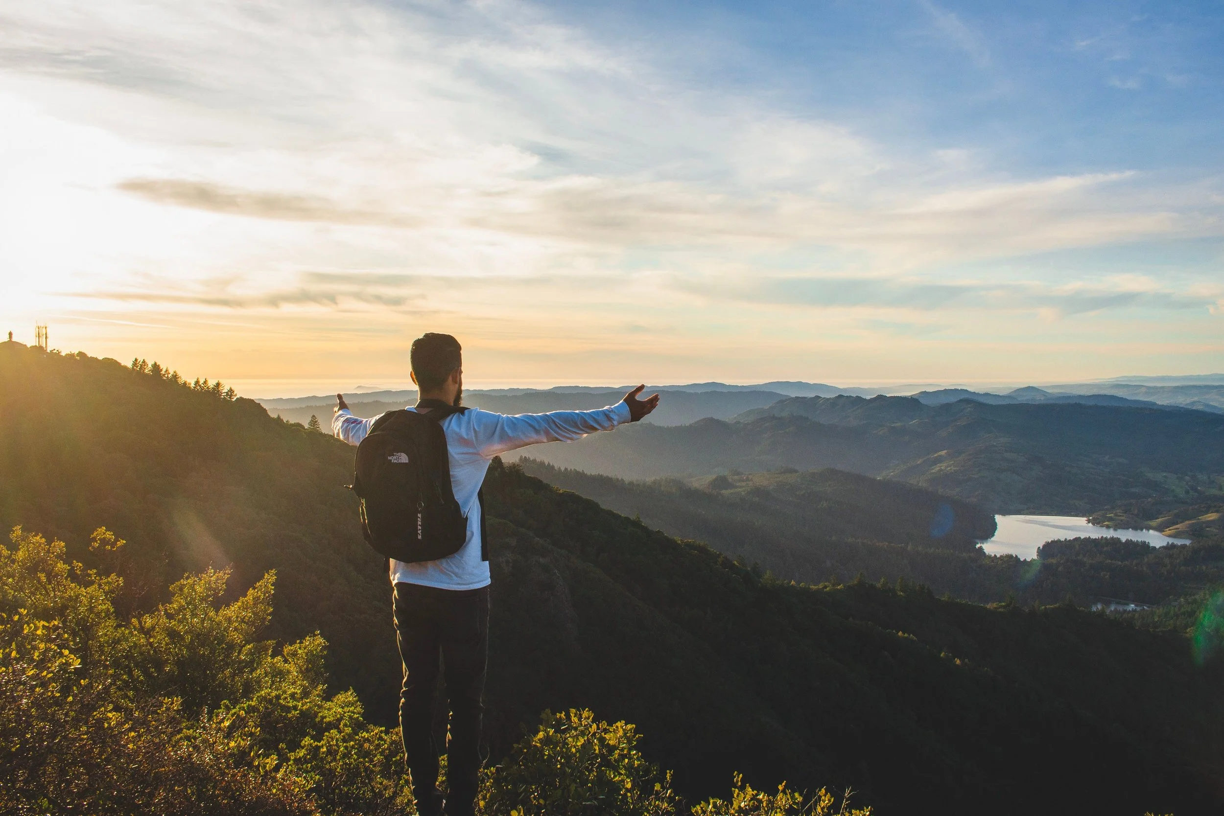 Man with a backpack standing on a hilltop with arms outstretched, overlooking mountains and a river at sunset.