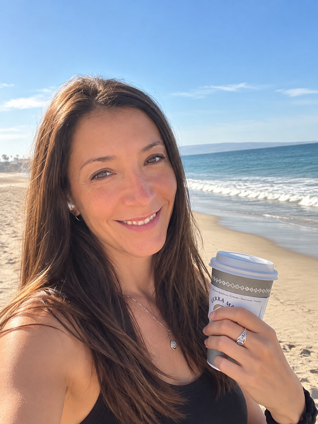 Emily Arden  smiling at the beach, holding a coffee cup, with the ocean and blue sky in the background.