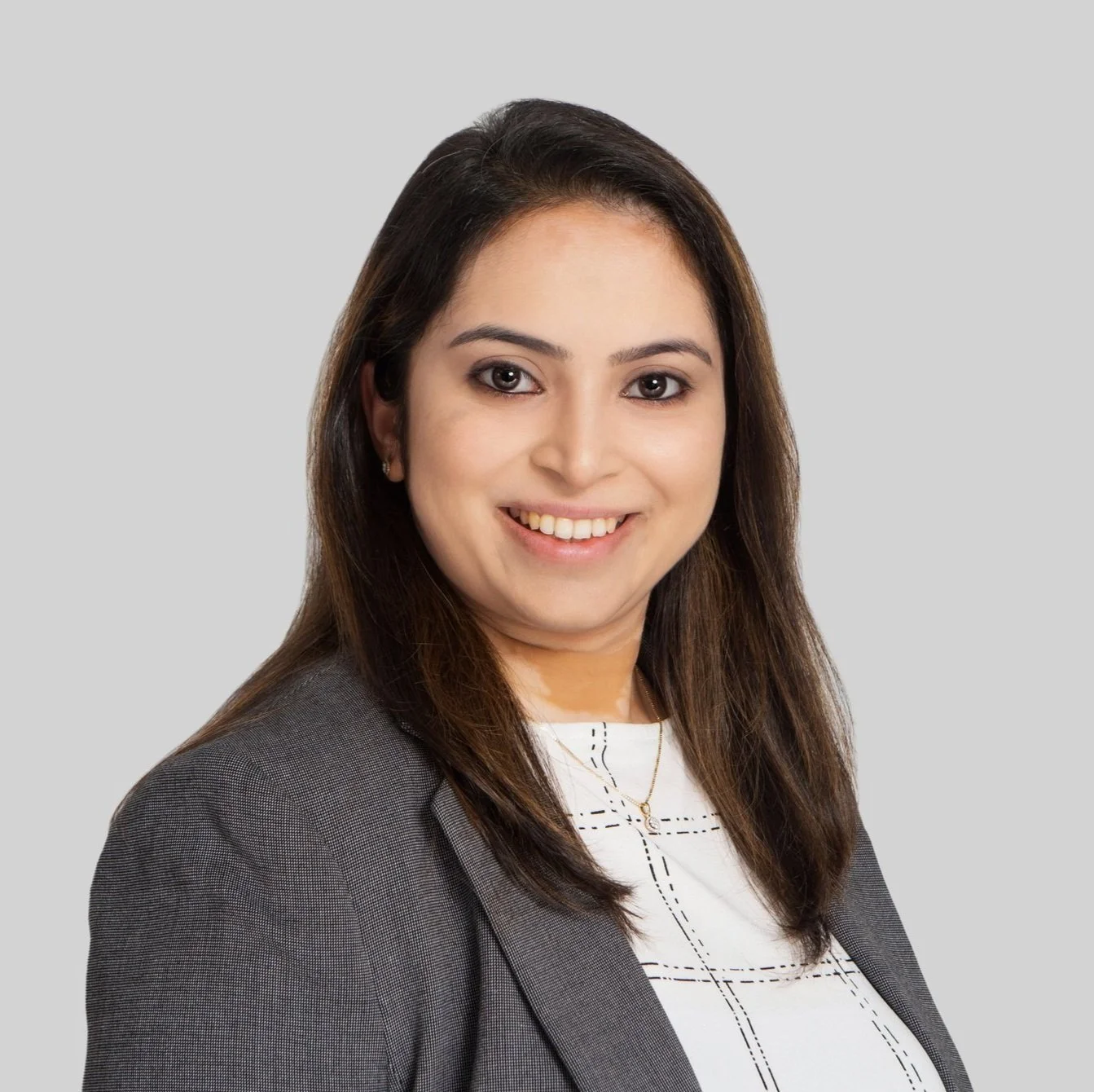 Professional headshot of a smiling woman with long brown hair, wearing a gray blazer over a white top with black stitching, against a light gray background.