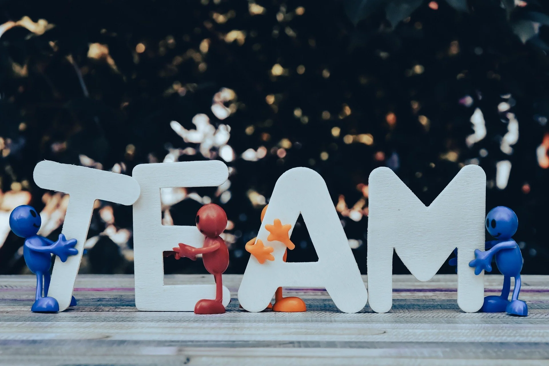 The word 'TEAM' spelled out with white wooden letters, with small colorful toy figures holding and interacting with the letters, on a wooden surface with blurry dark background.