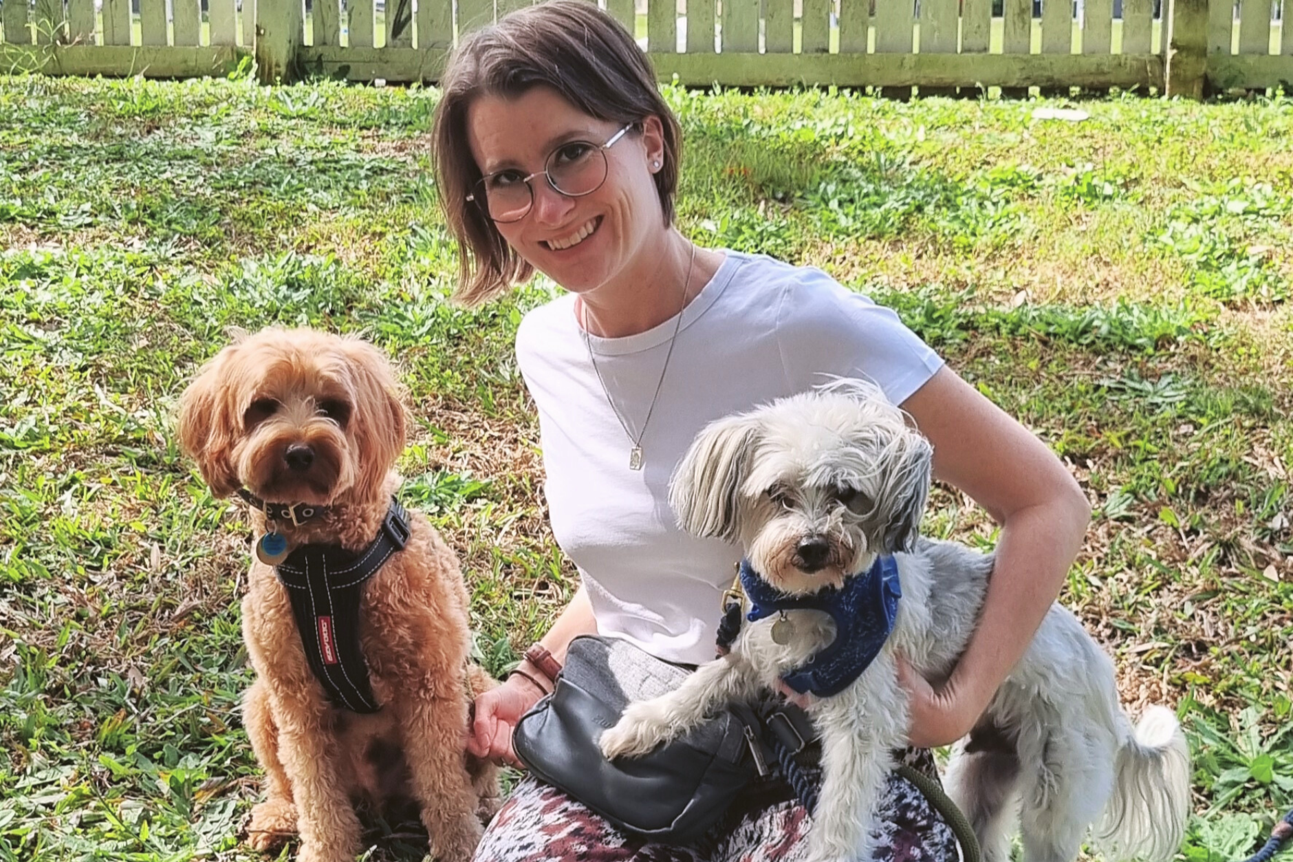 A woman with short brown hair, glasses, and a white t-shirt sitting in a grassy backyard with two dogs, one brown and one white, in a park-like setting with a wooden fence in the background.