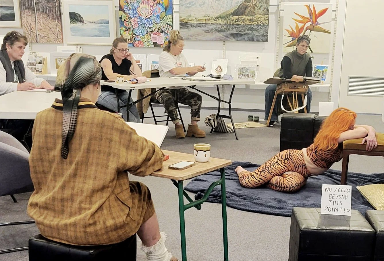 A woman with bright red hair dressed in a tiger-striped outfit is on her stomach, leaning on a piece of furniture, with her arms extended forward. Other women are seated at tables or standing in an art gallery or classroom, with paintings hanging on the walls behind them. There is a sign on a small black ottoman that reads 'NO ACCESS BEYOND THIS POINT!'