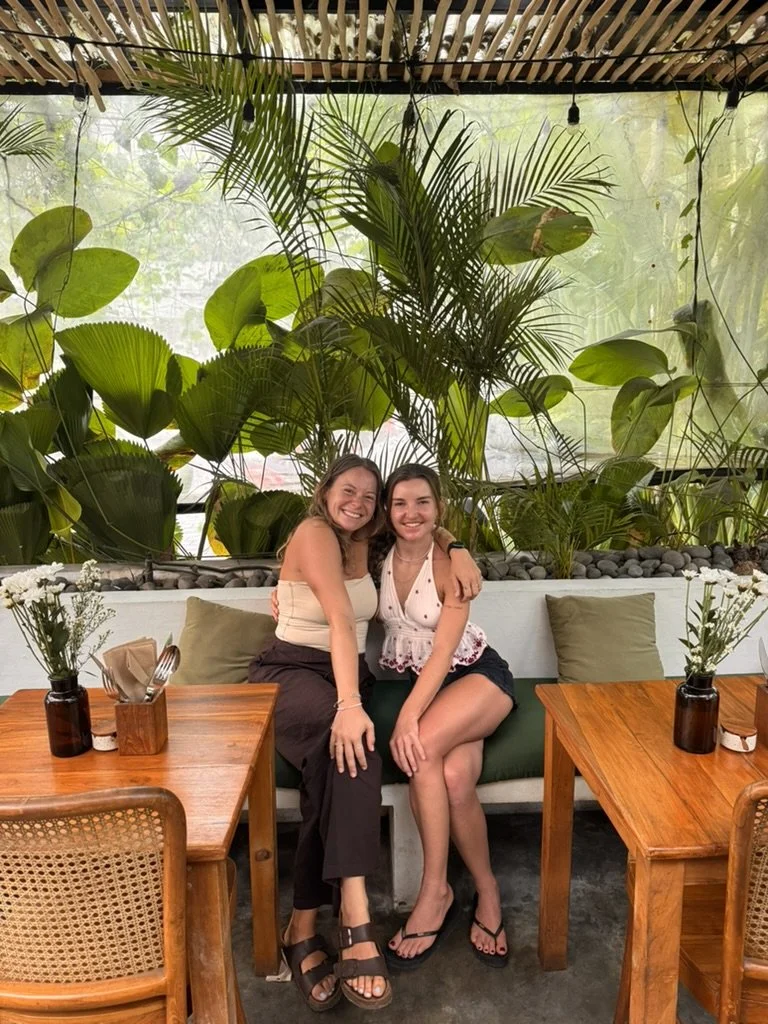 Two women sitting on a bench in a tropical-themed cafe, surrounded by large green plants. They are smiling and embracing each other, with wooden tables and chairs nearby, and small flower vases on the tables.