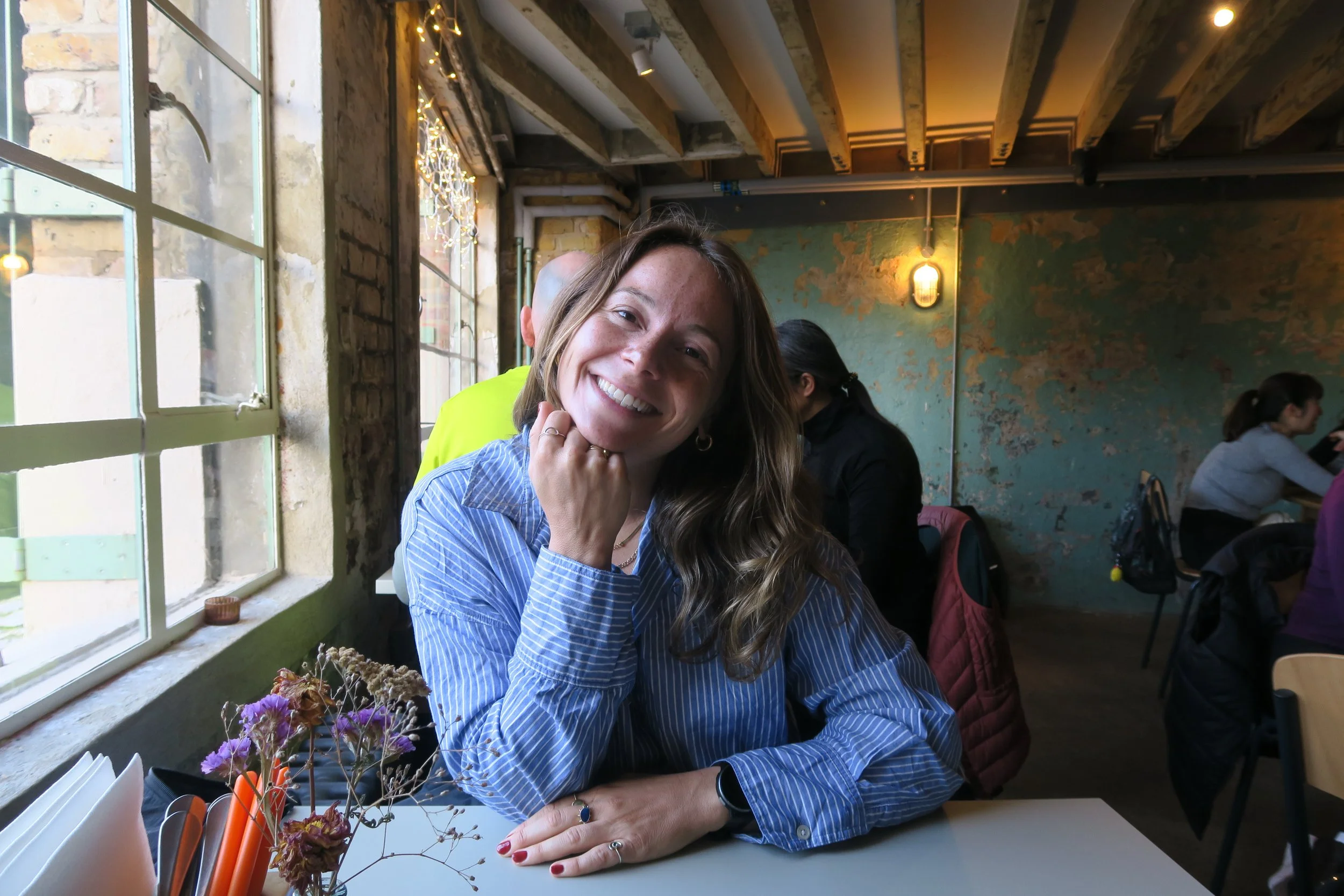 A woman with long wavy brown hair smiling at the camera while sitting at a table near a window in a cozy, rustic cafe. She is wearing a blue striped shirt, and her hand is resting under her chin. In front of her, there is a small bouquet of dried flowers and some notebooks or menus. The cafe has exposed brick walls, wooden beams, and warm lighting.