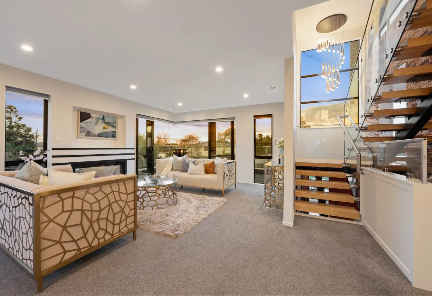 Modern living room with beige sofas, glass coffee tables, a plush area rug, large windows showing trees outside, and a staircase with wooden steps and glass railing.