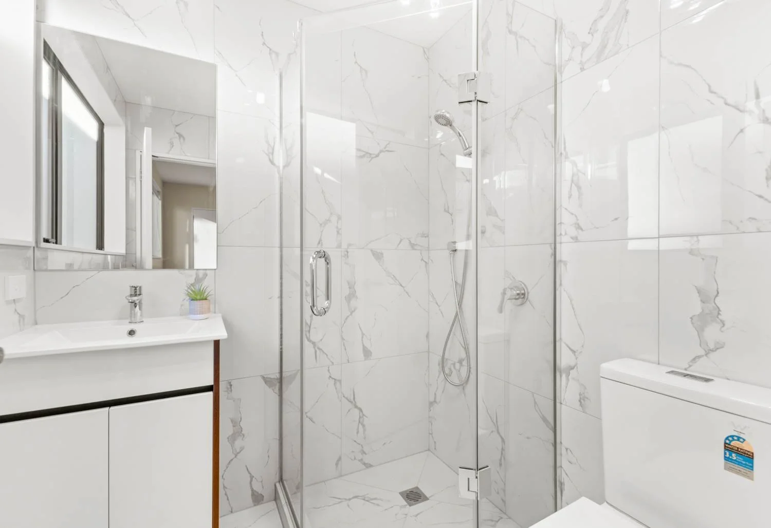 Modern bathroom with white marble walls and floor, glass shower enclosure with chrome handle, small white vanity with a plant on the countertop, mirror and a toilet with a blue sticker.