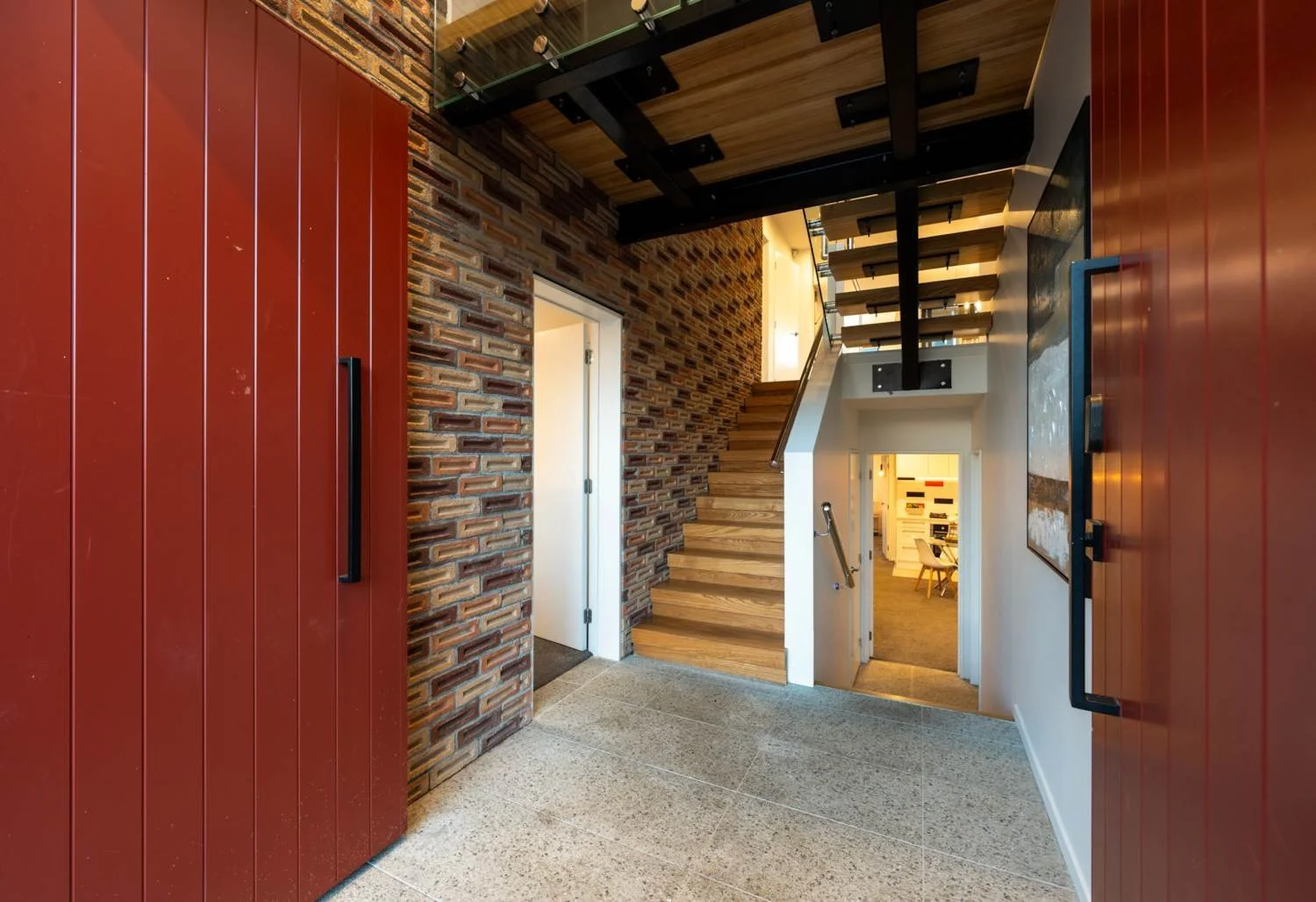 Interior house entryway with red front doors, brick wall on the left, wooden stairs leading up, and a doorway revealing a room with a dining table, chairs, and modern artwork.