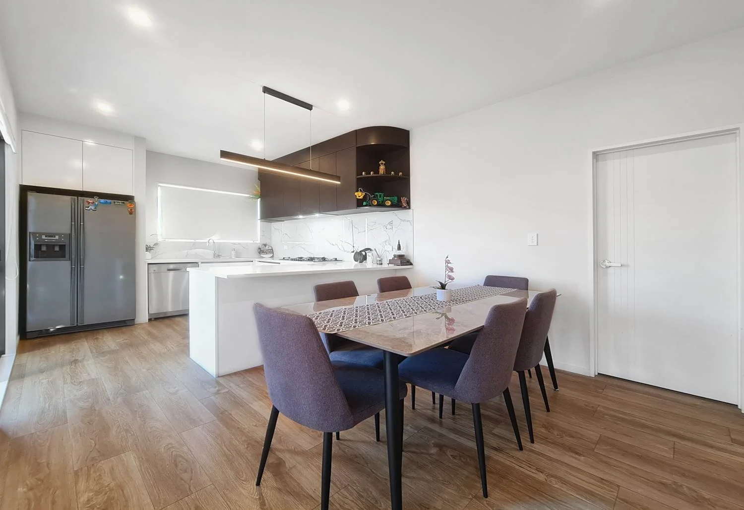 Modern kitchen and dining area with wooden floors, white walls, a brown dining table surrounded by six purple chairs, a white kitchen island, gray refrigerator, white sink, black cabinetry with open shelves containing decorative toys, and a window wi