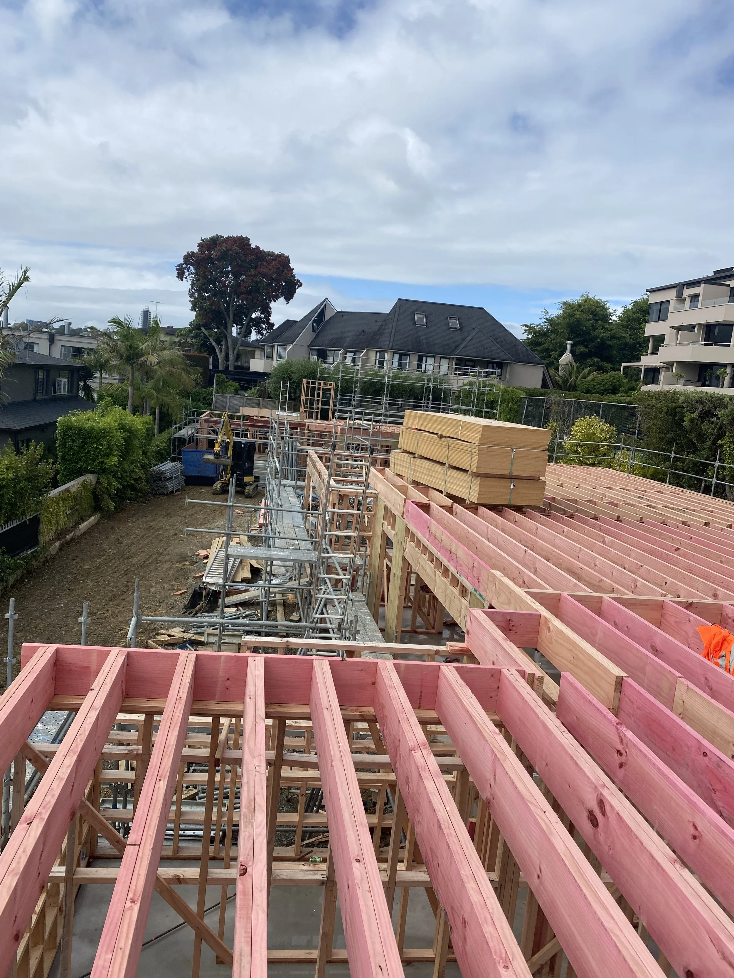 Construction site of a building with wooden framing and scaffolding, surrounded by residential houses and trees.