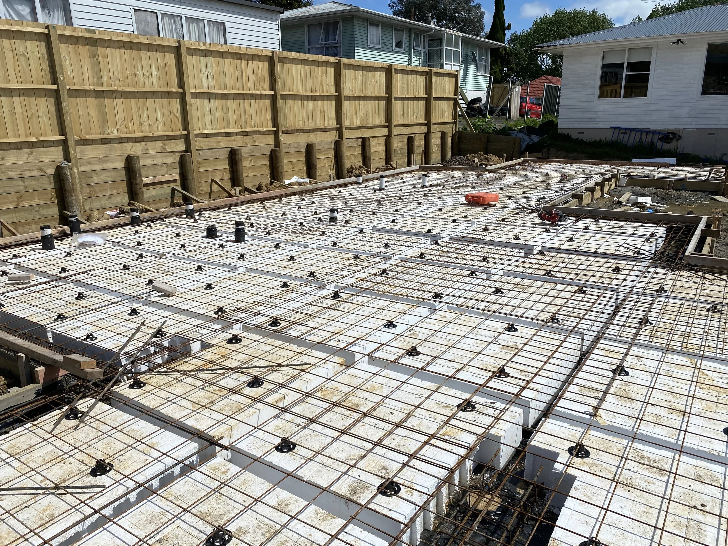 Construction site with rebar and foam blocks for a concrete foundation, surrounded by residential houses and a wooden fence.