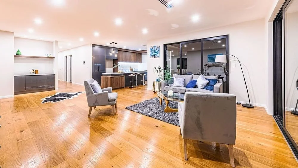 Open-concept living room with hardwood floors, a gray sofa with blue cushions, two gray armchairs, a glass coffee table, and a large sliding glass door leading outside. The space also includes a modern kitchen with dark cabinets and a breakfast bar.