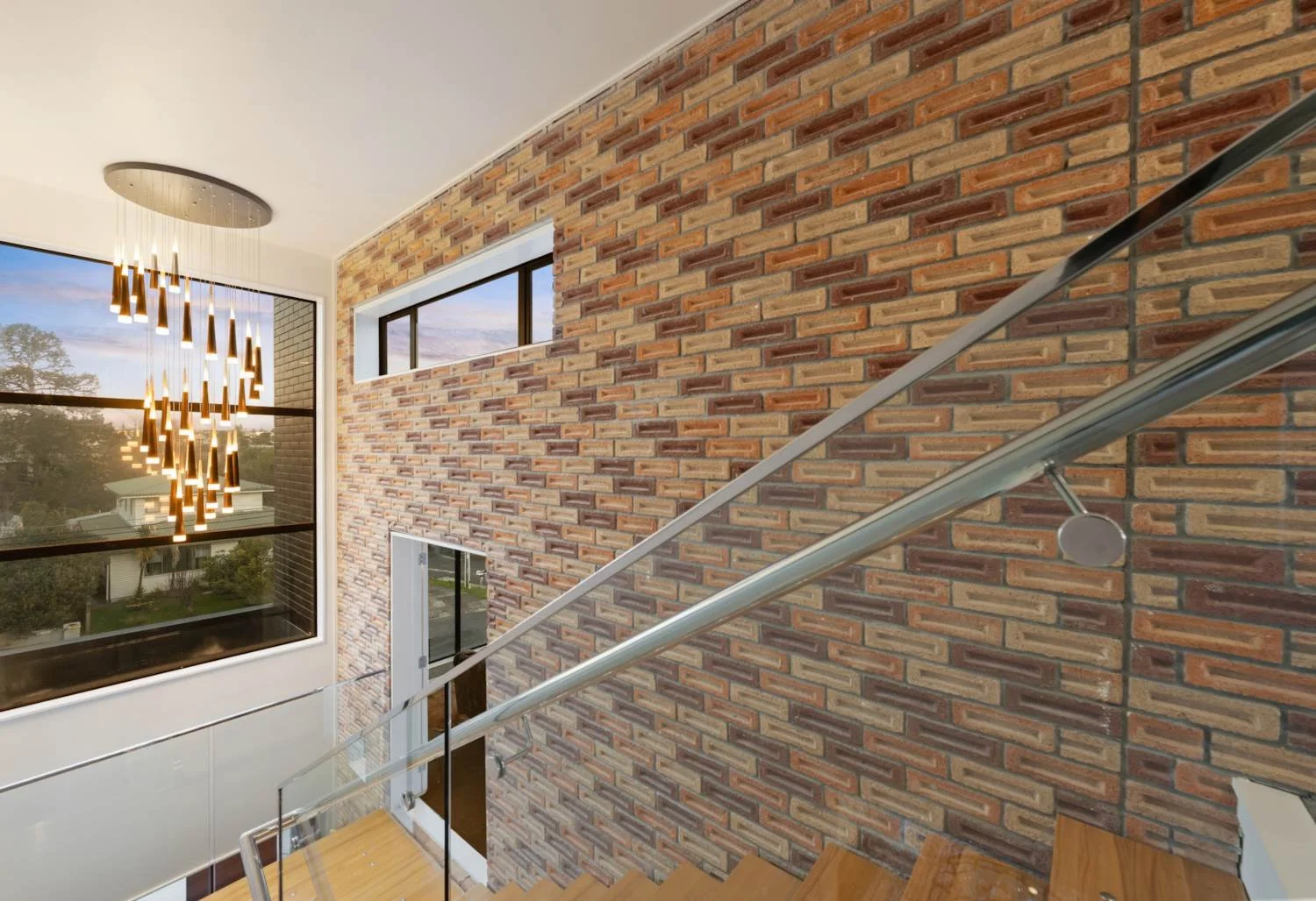Interior view of a modern house staircase with brick wall, large window, and hanging chandelier