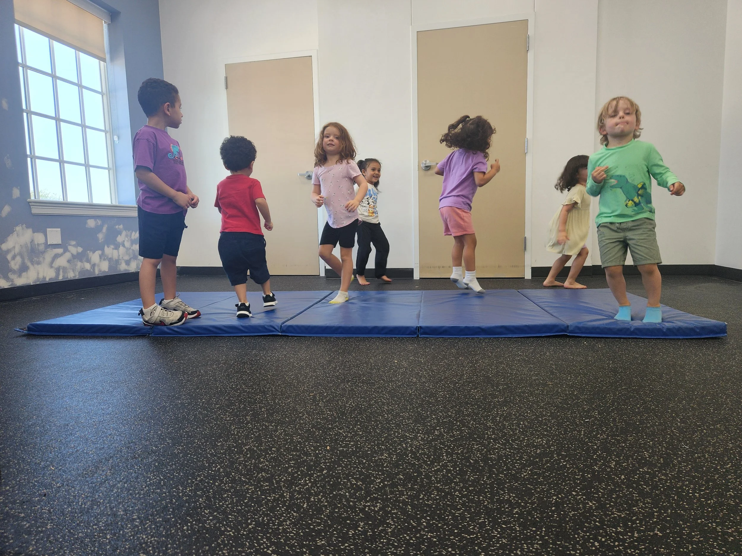 Children playing and jumping on a blue mat inside a room with a window and light-colored walls.